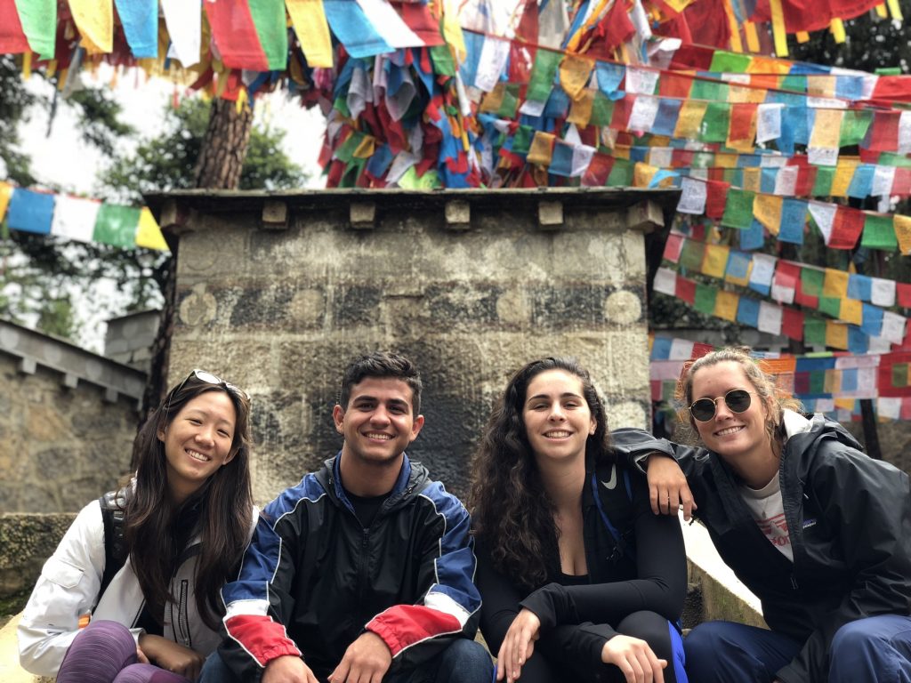 four students sitting behind stone monument and prayer flags
