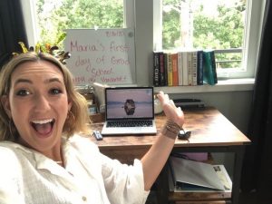 girl smiling in front of computer and desk and white board that celebrates first day of grad school online
